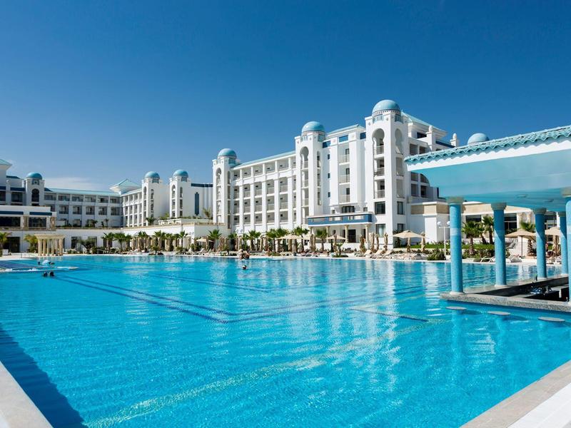 Large hotel with bright building and spacious clear outdoor pool under blue sky.