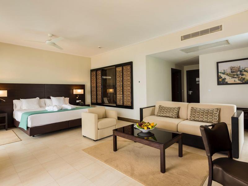 Modern furnished hotel room with bed, sofa, armchairs, and a table with flowers.