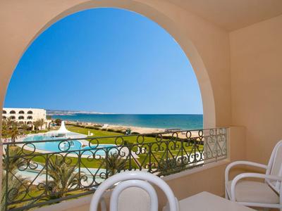 Blick von einem Balkon mit weißen Stühlen auf einen Pool und das Meer bei klarem Himmel.