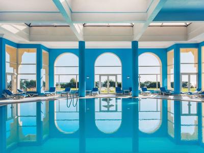 Innenraum eines Hallenbades mit blauen Säulen, großen Fenstern und klarem Wasser im Schwimmbecken.