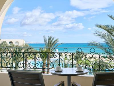 Balcón con mesa y sillas con vista a palmeras y al mar azul bajo un cielo despejado.