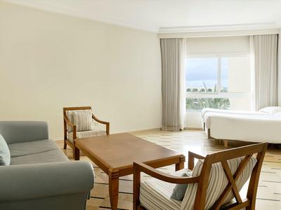 Helles Hotelzimmer mit Sofa, zwei Sesseln, Holztisch und Bett vor einem Fenster mit Vorhängen.