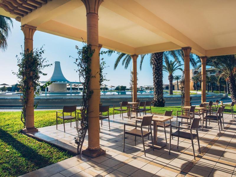 Sonnige Terrasse mit Tischen und Stühlen, Blick auf Palmen und Hotel am Meer.