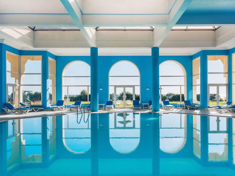 Innenraum eines Hallenbades mit blauen Säulen, großen Fenstern und klarem Wasser im Schwimmbecken.