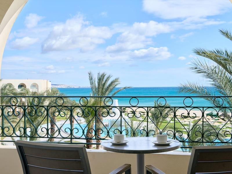 Balkon mit schwarzem Geländer, Tisch, Stühlen, Blick auf Palmen, Strand und blaues Meer unter blauem Himmel.
