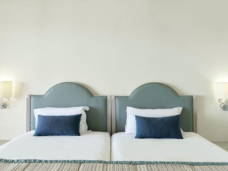 Zwei nebeneinanderstehende Betten mit grauen gepolsterten Kopfteilen, weißen Laken und blauen Kissen.