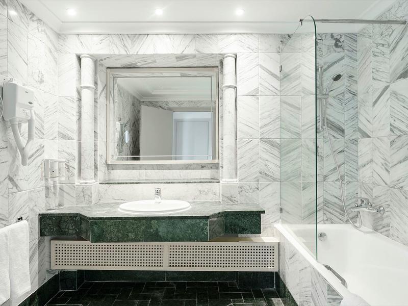 Helles Badezimmer mit weißen Marmorfliesen, großem Spiegel, schwarzer Waschtischplatte und Badewanne.