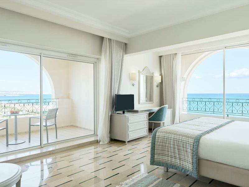 Helles Hotelzimmer mit Doppelbett und Blick auf Balkon mit Meerblick.