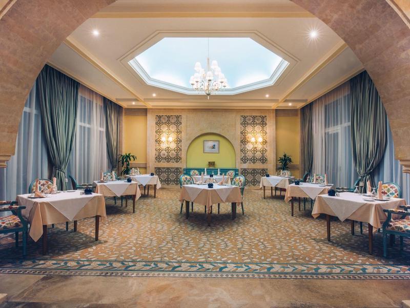 Eleganter Speisesaal mit runden und rechteckigen Tischen, blauen Vorhängen und Kronleuchter.