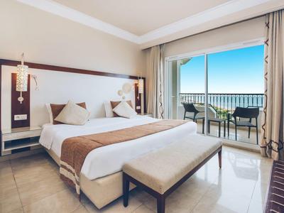 Modernes Hotelzimmer mit großem Doppelbett, Sitzbank, Balkon mit Meerblick und hellen Vorhängen.