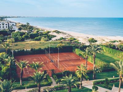 Tennisplatz mit roten Sand neben Palmen und Strand, dahinter blauer Himmel und Meer.