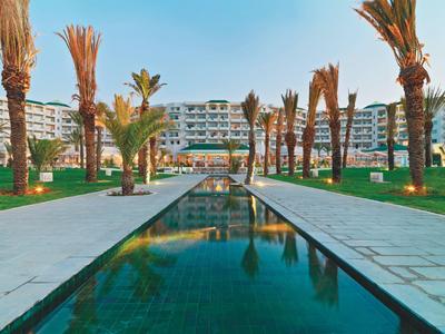Moderner Hotelkomplex mit langem Wasserbecken, Palmenreihen und klarem Himmel im Hintergrund.