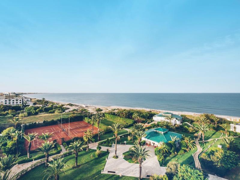 Blick auf ein Resort mit Tennisplätzen, Pool und Garten am Meer unter blauem Himmel.