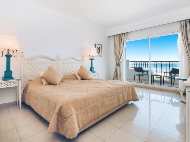 Helles Hotelzimmer mit Doppelbett, Nachttischen, Lampen und Balkon mit Meerblick.