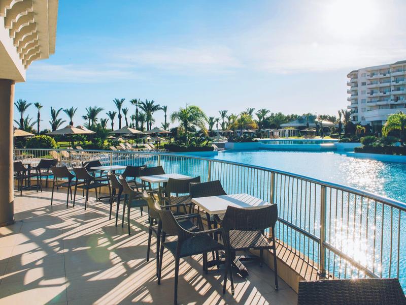 Sonnige Terrasse mit Tischen und Stühlen am Pool eines Hotels neben Palmen und modernen Gebäuden.