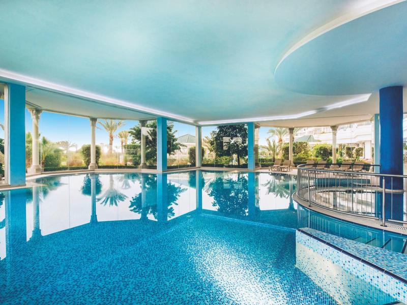 Hallenbad mit großen Fenstern, blauem Wasser und überdachtem Bereich mit Aussicht auf Palmen