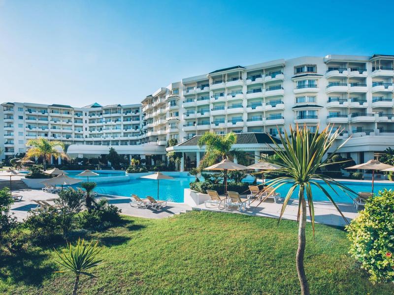 Großes, modernes Hotel mit Außenpool und Liegestühlen, umgeben von Grünanlagen.