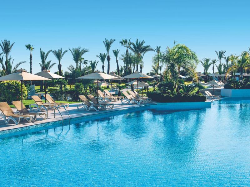 Großer Hotelpool mit Liegestühlen, Sonnenschirmen und Palmen unter klarem blauem Himmel.