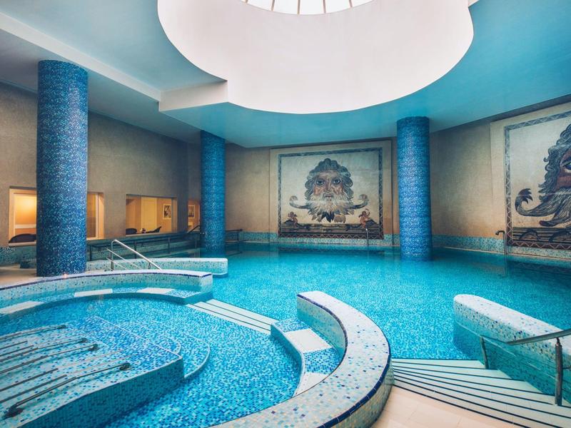 Innenpool mit blauen Säulen, Rundfenster und Mosaikwänden in einem Hotel.