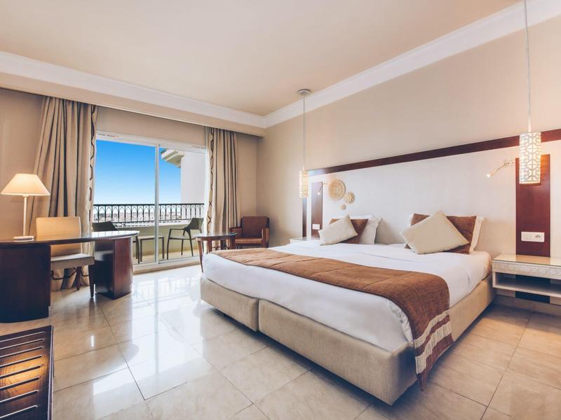 Hell eingerichtetes Hotelzimmer mit großem Bett und Balkon mit Meerblick.