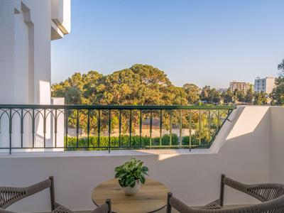Balcony with table and chairs overlooking green trees and buildings under clear sky.