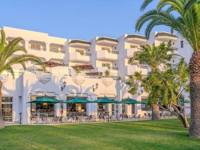 Hotel building with balconies, green lawn, and palm trees in front under sunny weather.