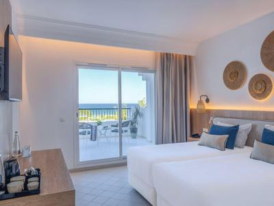 Bright hotel room with two beds and view of terrace and sea.