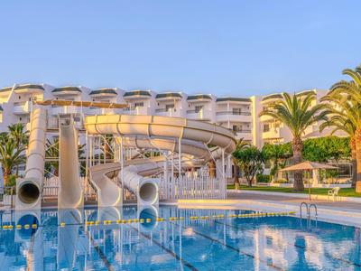 Hotel pool with water slides, surrounded by palm trees and a white hotel building.