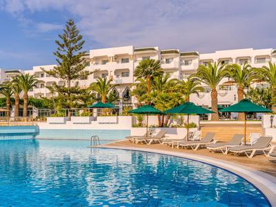 Hotel with large pool, sun umbrellas and lounge chairs surrounded by palm trees under blue sky.