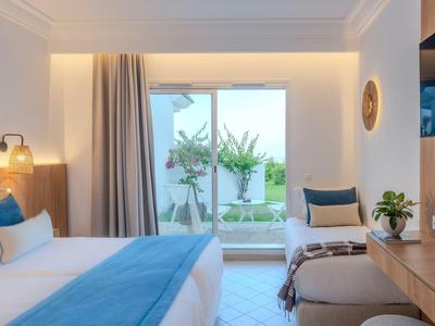 Bright hotel room with double bed, blue and white bedding, window overlooking a garden.