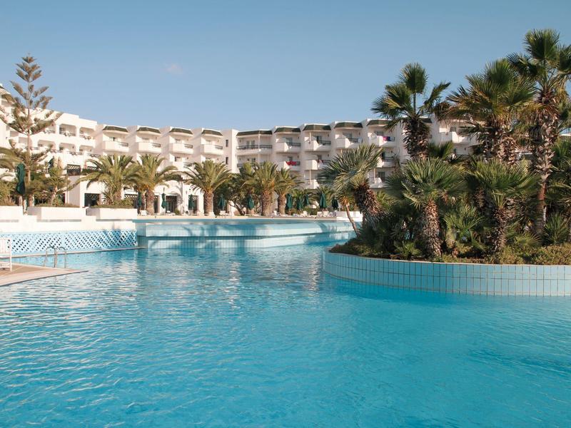 Large hotel pool with palm trees and modern accommodation in the background under clear sky.