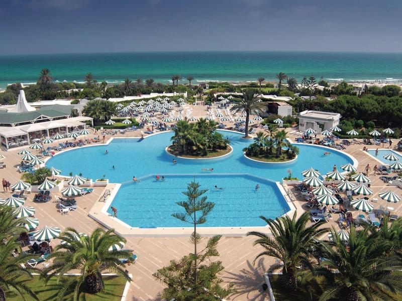 Large hotel pool with sun umbrellas and palm trees by the sea.
