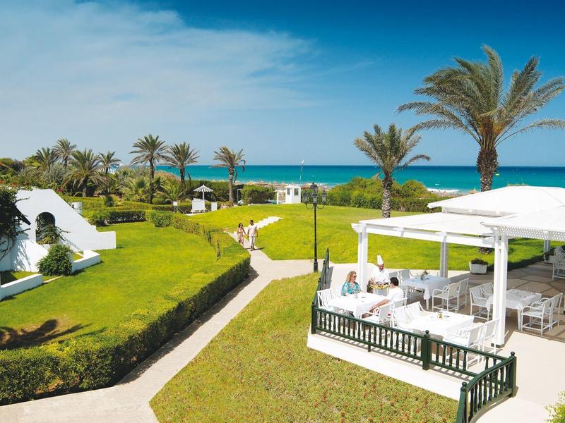 Hotel garden with pathways, palm trees, a white pavilion, and a view of the blue sea under a sunny sky.