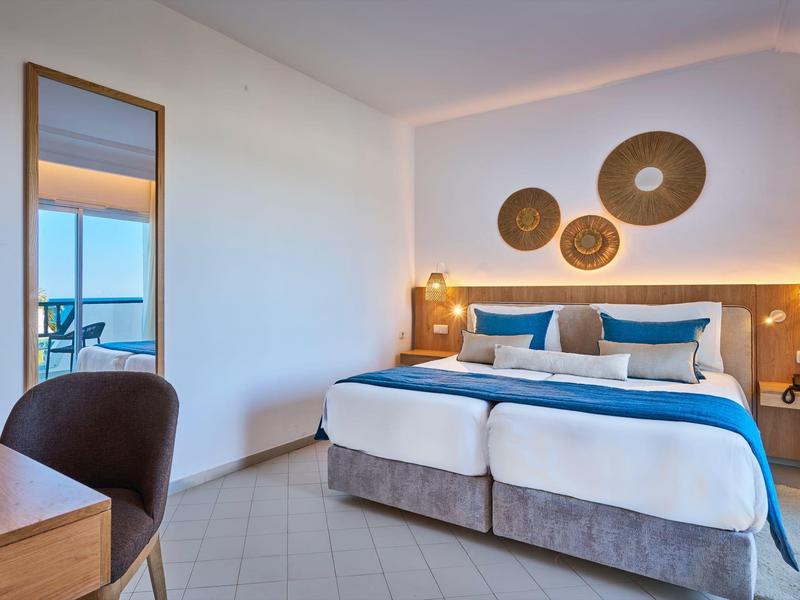 Modern hotel room with double bed, blue accents, and view of balcony and sea.