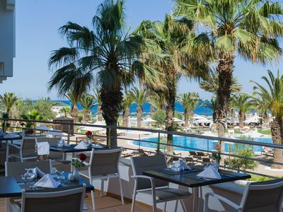 View of a terrace with dining tables and chairs, palm trees, and a pool by the sea in the background.
