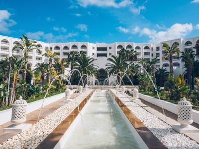 Luxe hotelgebouw met weelderige palmbomen en een lang waterbassin op de voorgrond.