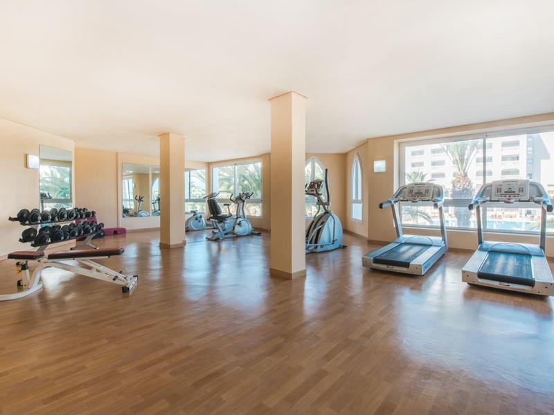 Bright gym with wooden floor, dumbbells, treadmills, and fitness equipment by windows.
