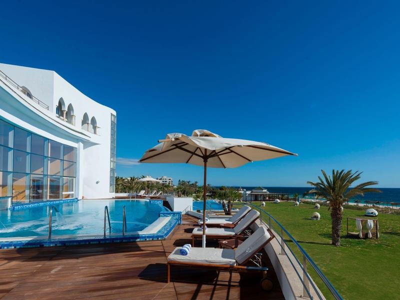 Modern hotel pool with sun loungers and umbrella overlooking the sea.