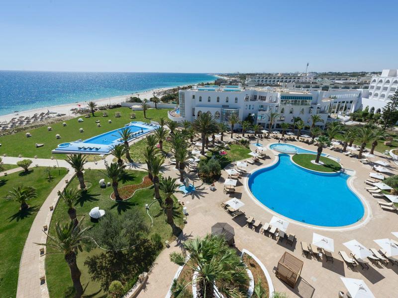 Large hotel complex with pool, sun loungers, and view of a long sandy beach.