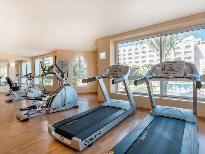 Fitness room with various exercise machines and large windows overlooking a hotel and palm trees.