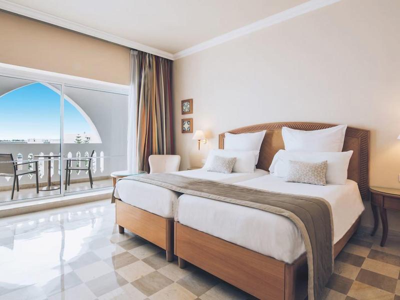 Bright hotel room with two single beds and balcony overlooking the sea.