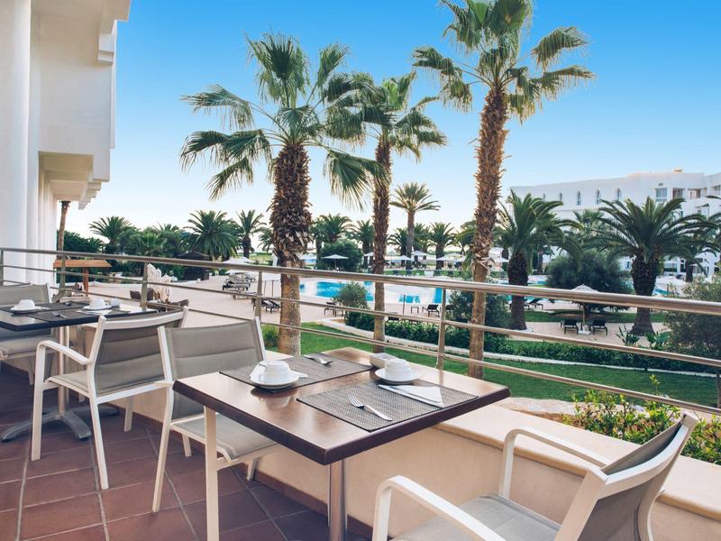 Balcony with tables and chairs overlooking palm trees and a pool.