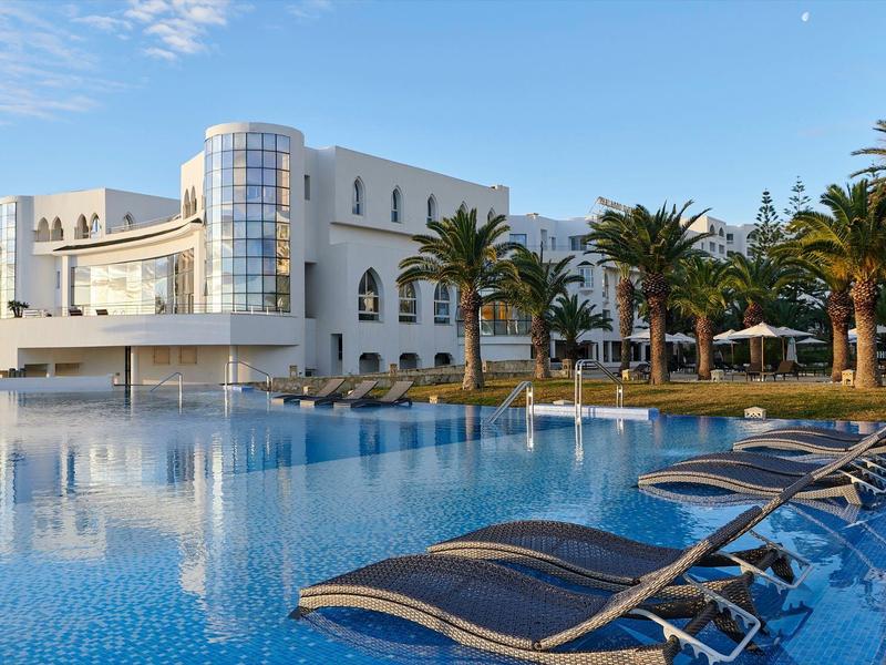 Modern hotel complex with pool, loungers, and palm trees under clear sky.