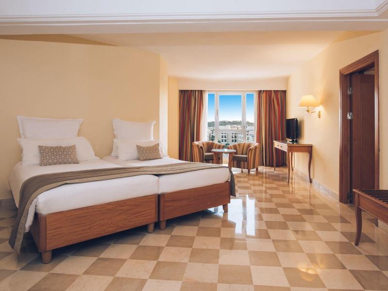 Bright hotel room with double bed, chairs, and sea view through large windows.