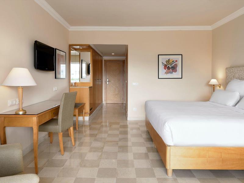 Bright hotel room with double bed, desk with chair, wall-mounted TV, and neutral tones.