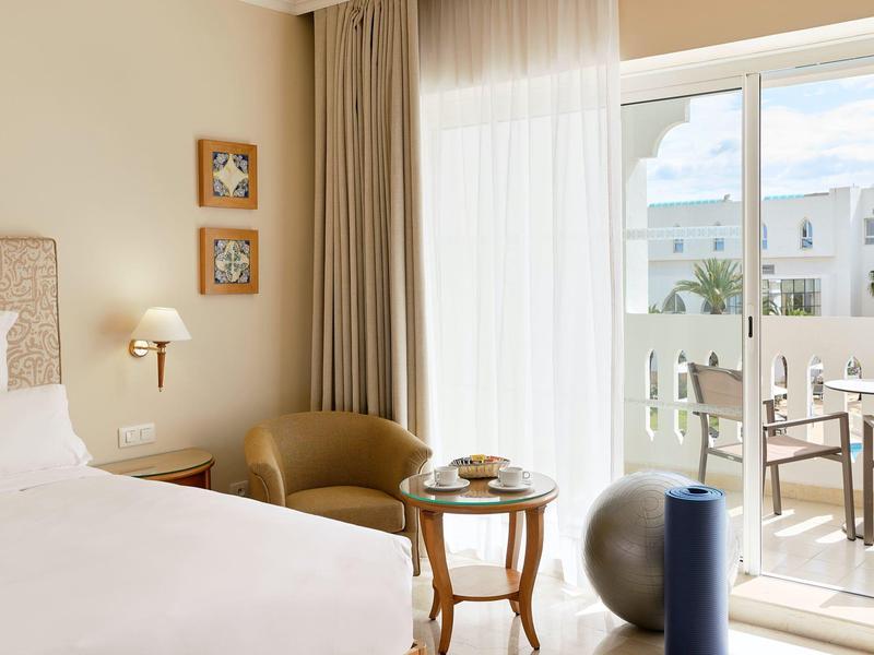 Bright hotel room with bed, armchair, table, and balcony access overlooking neighboring buildings.