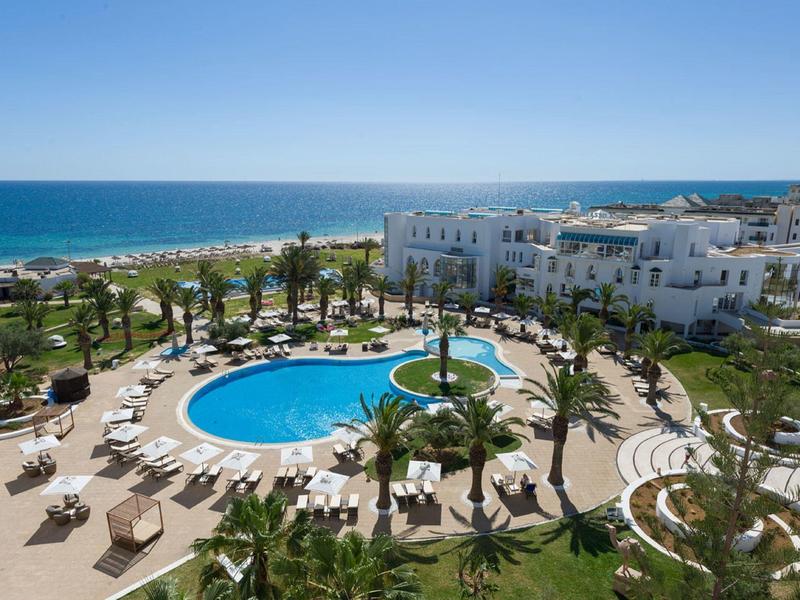 Hotel complex with circular pool, lounge chairs, and sea view under clear sky.