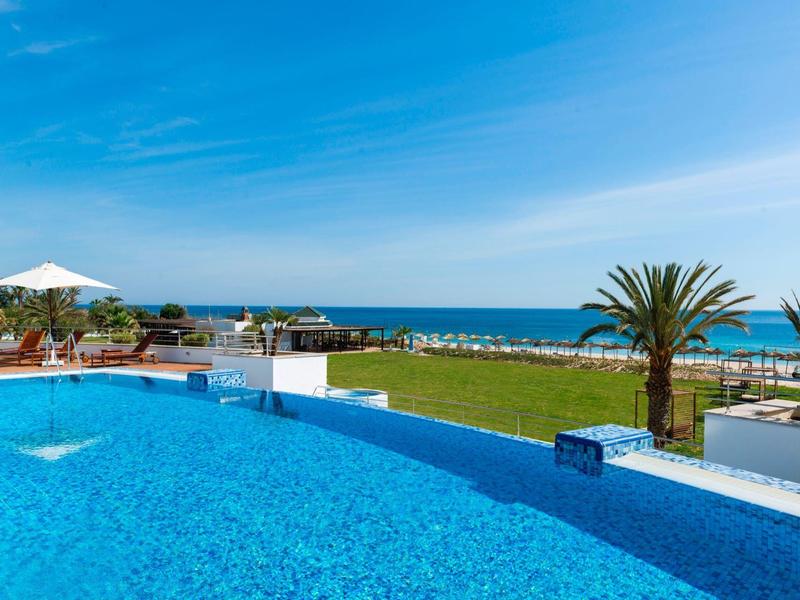 Infinity pool overlooking the sea, surrounded by palm trees and sun loungers under a blue sky.