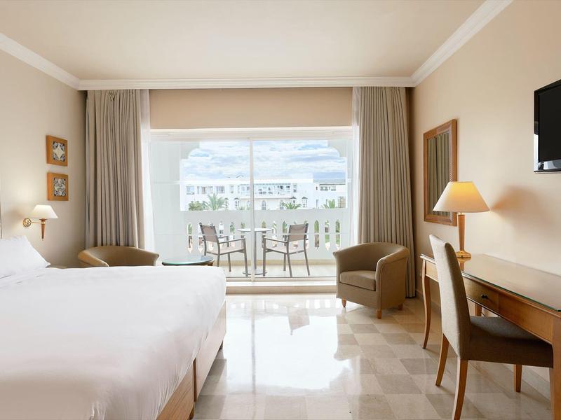 Hotel room with large bed, desk, TV, and balcony with seating and sea view.