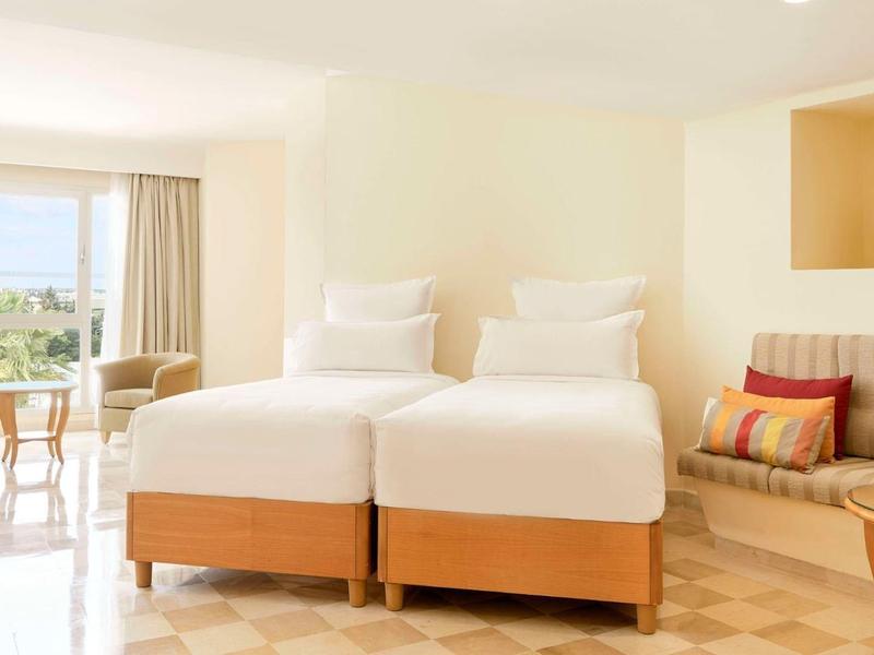 Bright hotel room with two single beds, seating area, and window view.